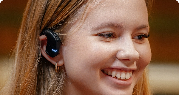 Et nærbillede af en smilende ung kvinde, der bærer en sort Vasco Translation E1-øretelefon. Billedet fremhæver enhedens hygiejniske, åbne design, der viser, hvordan den sidder behageligt uden på øret uden at trænge ind i øregangen. 