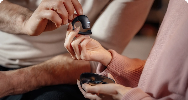 Et nærbillede af en kvindes hænder, der forsigtigt holder en sort Vasco Translation E1-øretelefon mellem fingerspidserne, hvilket fremhæver dens lette design. En mands hånd rører ved enheden ovenfra, mens hun i den anden hånd holder et åbent, sort opladningsetui. 