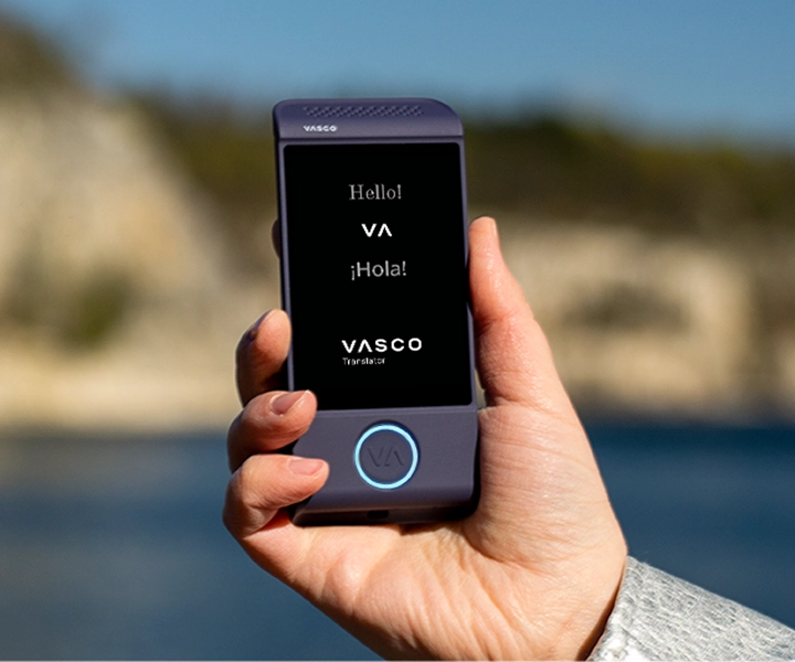 Et nærbillede af en hånd, der holder en lilla Vasco Translator M4 elektronisk oversætter mod en lys udendørs baggrund. Skærmen viser hilsenerne »Hello!«, »¡Hola!« og »VASCO Translator«-logoet.