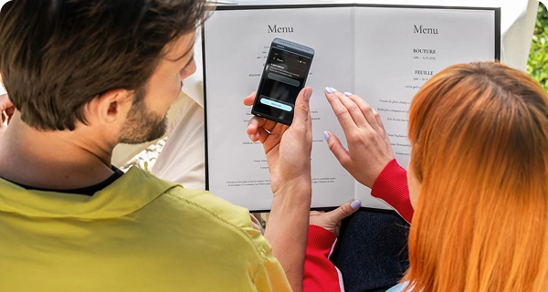 Et billede set over skulderen af en mand og en kvinde med lysorange hår, der sidder ved et bord på en restaurant og kigger på en menu. Manden holder en sort Vasco Translator Q1, hvor der vises en »Del«-skærm med et indtastningsfelt til at sende oversættelsen via e-mail. I baggrunden ses en hvid dug og restaurantens indretning.
